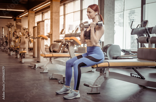 Red-haired fitness woman sitting on a bench with dumbbells in the gym