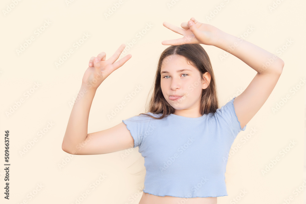 Young Caucasian girl making funny gesture with tongue out and fingers in victory sign on beige isolated background Young Caucasian girl making funny gesture with tongue out and fingers in victory sign on beige isolated background