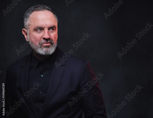 Studio shot of stylish aged man with beard dressed in black coat and black shirt.
