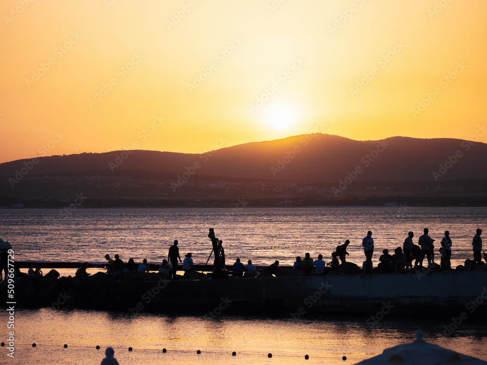 Silhouettes of many people on pier in sea bay against backdrop of sunset and mountains. Summertime and relax concept with copy space.
