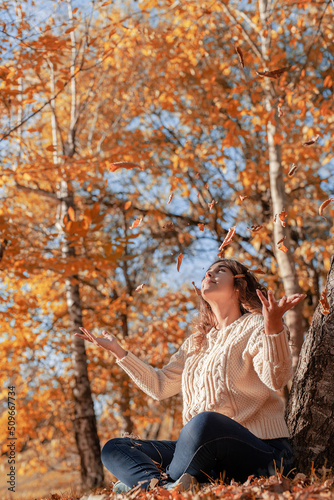 Young woman throwing yellow leaves up in the air sitting in the autumn forest