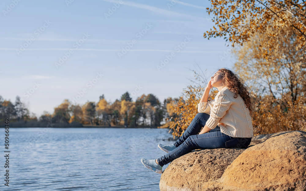 Thoughtful romantic woman sitting on rocks on the river bank in sunset in autumn day