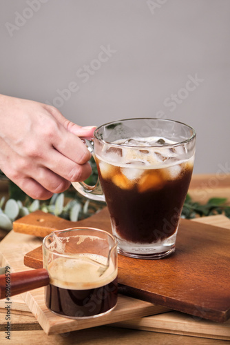 woman used hand take cold iced black brew coffee in a glass with pieces of ice on a wooden table, natural light, fresh summer drink in the morning