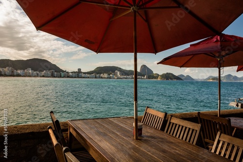 Fototapeta Naklejka Na Ścianę i Meble -  Rio de Janeiro, Brazil. Beach and sea and buildings on the edge of Copacabana Bay, mountains and Sugar Loaf Mountain seen from Copacabana Fort. Wooden tables and chairs with umbrellas.
