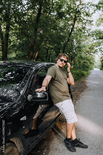 Stylish long-haired hippie man in sunglasses stands near a black car on the road. Photography, travel, business. © shchus