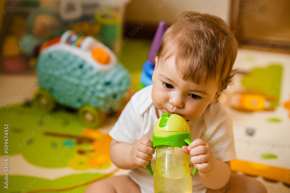 Cute little boy drinking water from a training bottle with a straw, home interior, lifestyle Cute little boy drinking water from a training bottle with a straw, home interior, lifestyle
