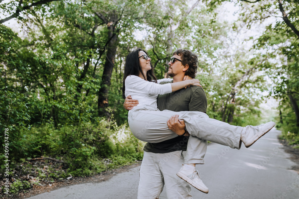 A bearded long-haired stylish man holds his beloved hippie brunette woman in sunglasses in his arms and circles, dances her in nature in a park on the road. Portrait, photograph of smiling newlyweds. A bearded long-haired stylish man holds his beloved hippie brunette woman in sunglasses in his arms and circles, dances her in nature in a park on the road. Portrait, photograph of smiling newlyweds.