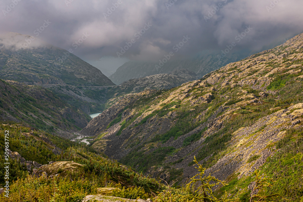 Naklejka premium Skagway, Alaska, USA - July 20, 2011: Klondike highway to Canada. Down the road, the valley with small pond under heavy cloudscape over green mountain flanks.