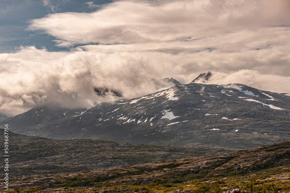 Fototapeta premium Skagway, Alaska, USA - July 20, 2011: Klondike highway to Canada. Thick cloudscape invades Canadian Rockies mountain with snow patches behind dry high desert with some green vegetation.