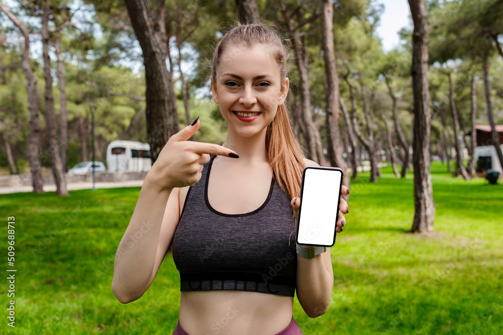 Young woman smiling confident wearing sports bra standing on city park, outdoors pointing finger at a mobile phone with blank white screen. Fitness app. Recommending new mobile application, mockup. Young woman smiling confident wearing sports bra standing on city park, outdoors pointing finger at a mobile phone with blank white screen. Fitness app. Recommending new mobile application, mockup.