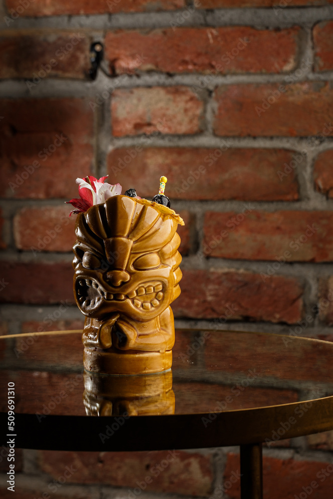 Tropical cocktail - zombie, served in a tiki style with fruits and flowers. An exotic cocktail in a tiki glass stands on a wooden bar counter in a pub or restaurant, space for text.
