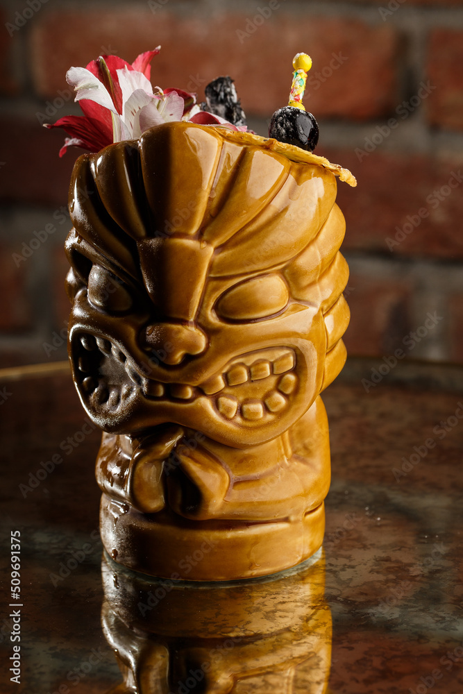 Tropical cocktail - zombie, served in a tiki style with fruits and flowers. An exotic cocktail in a tiki glass stands on a wooden bar counter in a pub or restaurant, space for text.