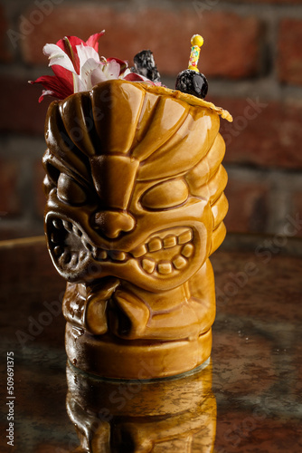 Obraz na płótnie Tropical cocktail - zombie, served in a tiki style with fruits and flowers. An exotic cocktail in a tiki glass stands on a wooden bar counter in a pub or restaurant, space for text. w salonie