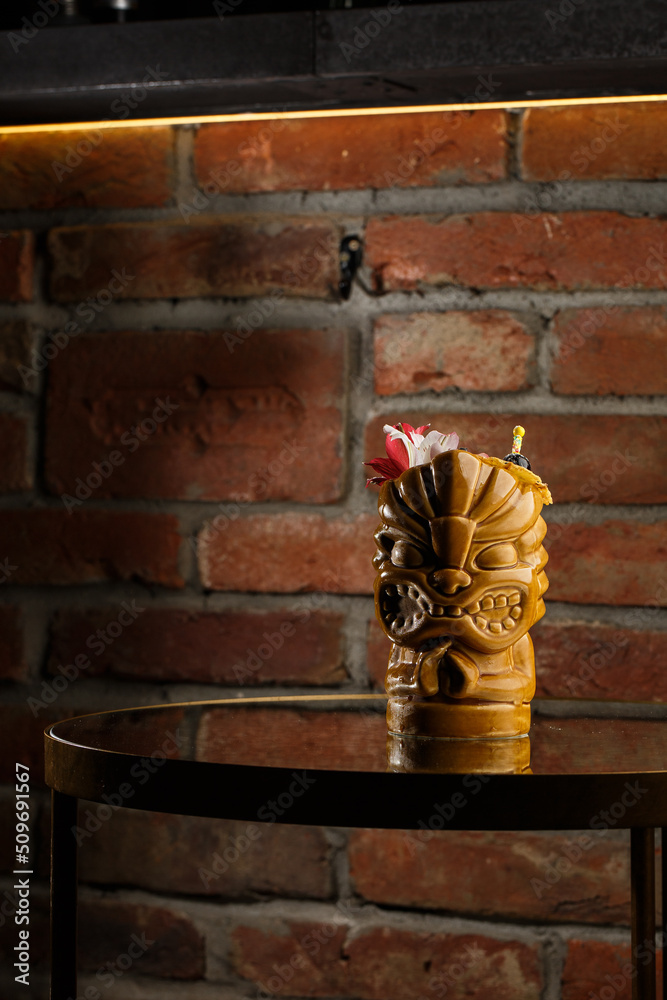 Tropical cocktail - zombie, served in a tiki style with fruits and flowers. An exotic cocktail in a tiki glass stands on a wooden bar counter in a pub or restaurant, space for text.