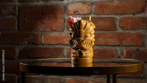 Obraz na płótnie Tropical cocktail - zombie, served in a tiki style with fruits and flowers. An exotic cocktail in a tiki glass stands on a wooden bar counter in a pub or restaurant, space for text. w salonie