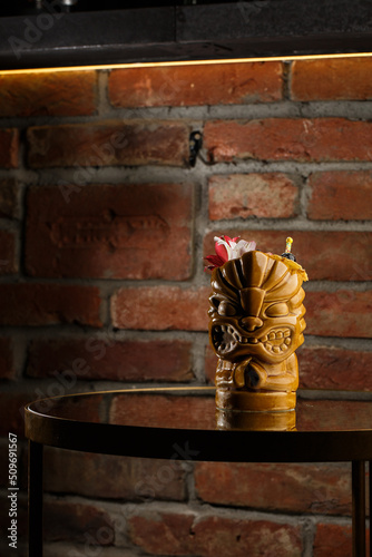 Obraz na płótnie Tropical cocktail - zombie, served in a tiki style with fruits and flowers. An exotic cocktail in a tiki glass stands on a wooden bar counter in a pub or restaurant, space for text. w salonie
