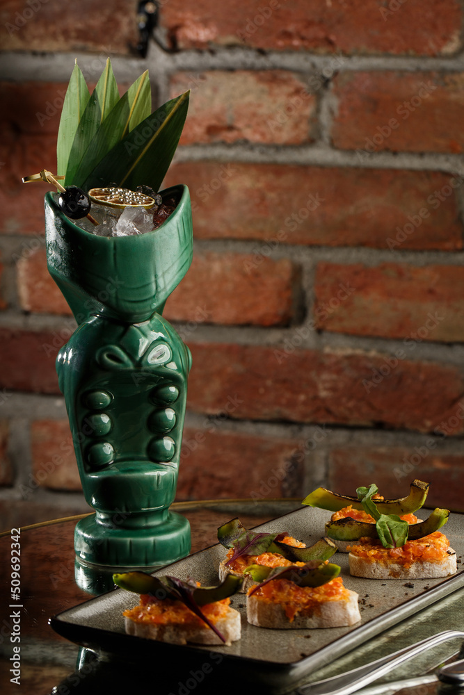 Tropical cocktail - zombie, served in a tiki style with fruits and flowers. An exotic cocktail in a tiki glass stands on a wooden bar counter in a pub or restaurant, space for text.