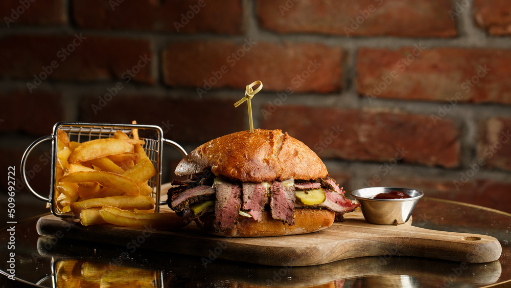Pastrami sandwich on rye bread served with pickles and mustard sauce. Tasty sandwich with french fries potatoes on dark background.