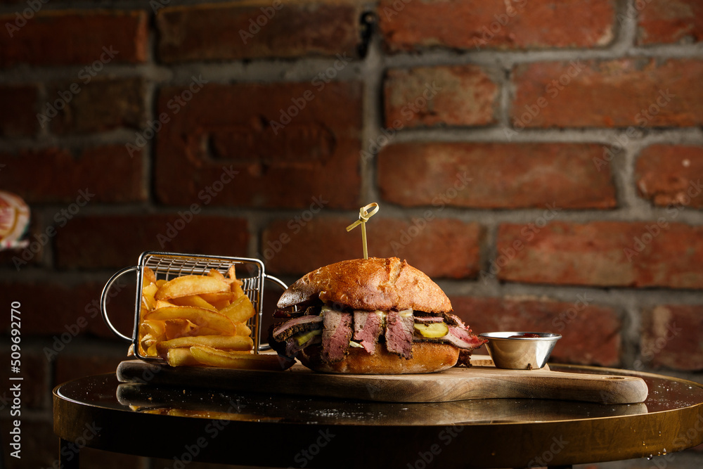 Pastrami sandwich on rye bread served with pickles and mustard sauce. Tasty sandwich with french fries potatoes on dark background.