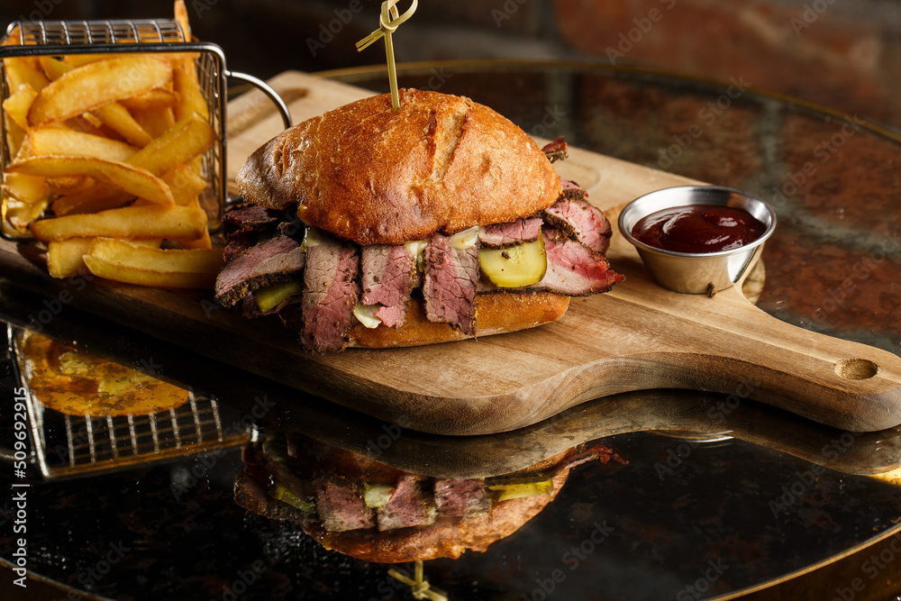 Pastrami sandwich on rye bread served with pickles and mustard sauce. Tasty sandwich with french fries potatoes on dark background.