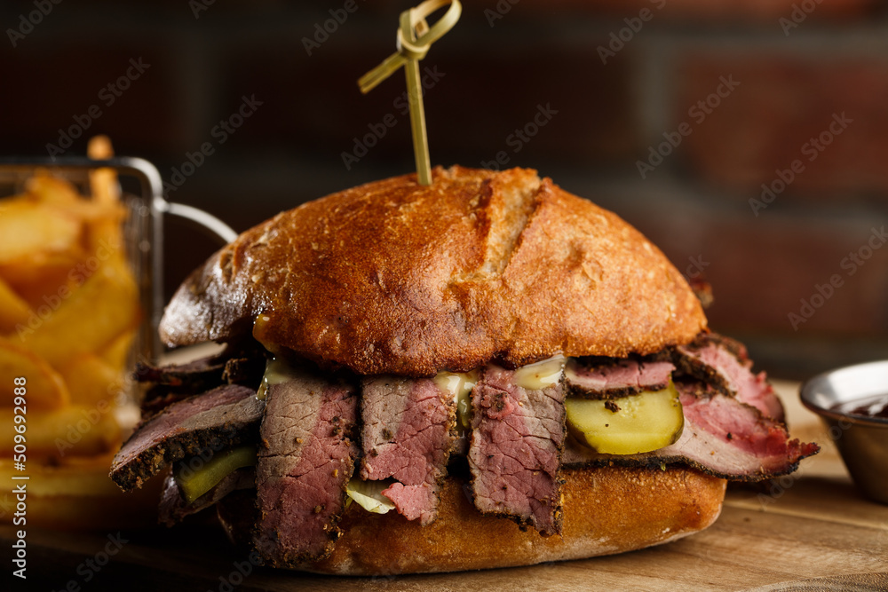 Pastrami sandwich on rye bread served with pickles and mustard sauce. Tasty sandwich with french fries potatoes on dark background.