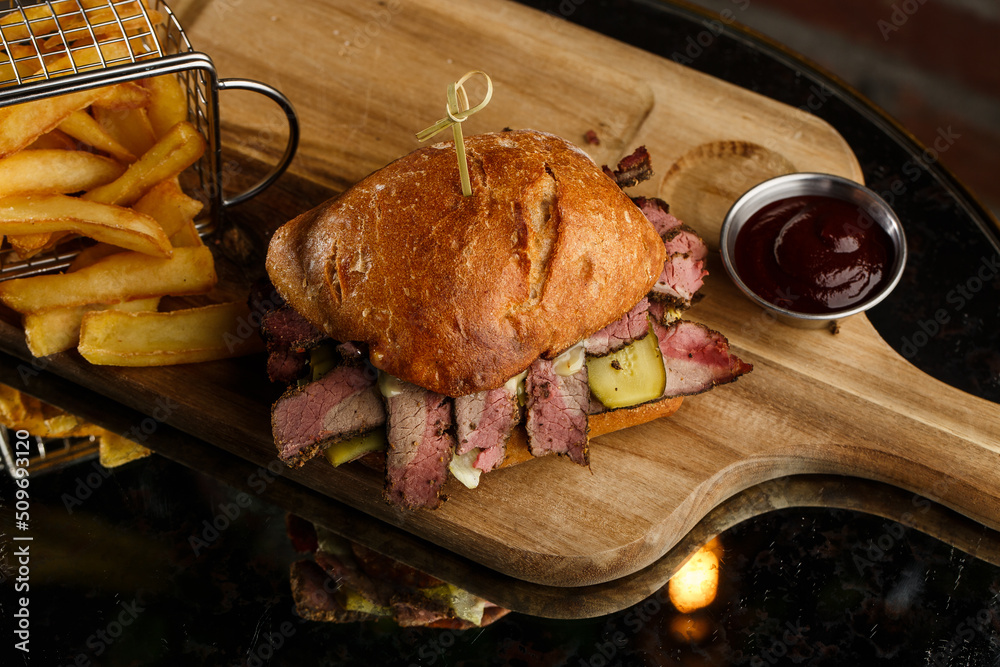 Pastrami sandwich on rye bread served with pickles and mustard sauce. Tasty sandwich with french fries potatoes on dark background.
