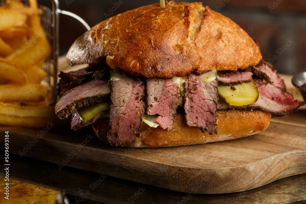 Pastrami sandwich on rye bread served with pickles and mustard sauce. Tasty sandwich with french fries potatoes on dark background.