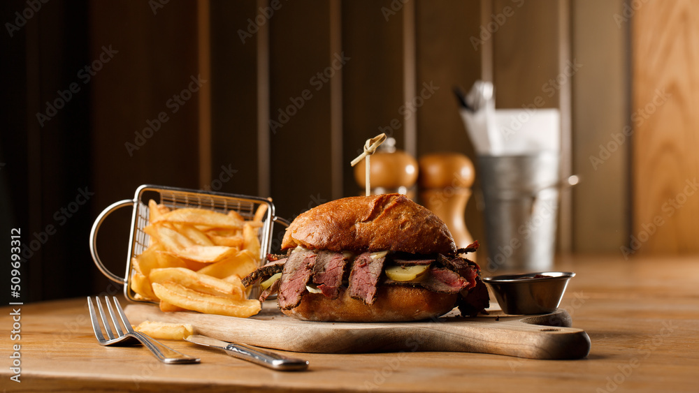 Pastrami sandwich on rye bread served with pickles and mustard sauce. Tasty sandwich with french fries potatoes on dark background.