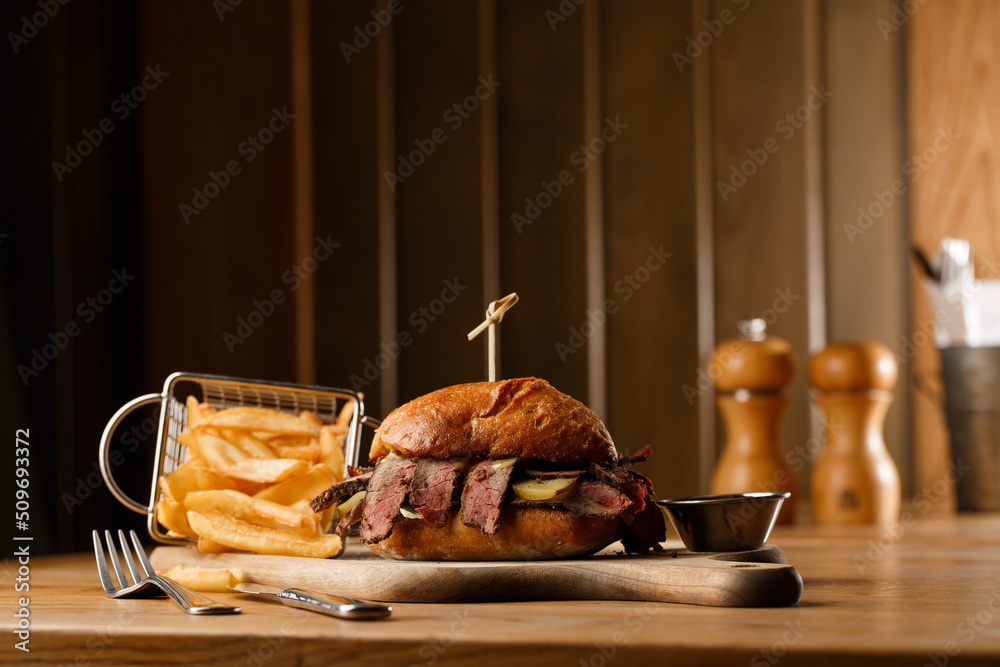 Pastrami sandwich on rye bread served with pickles and mustard sauce. Tasty sandwich with french fries potatoes on dark background.