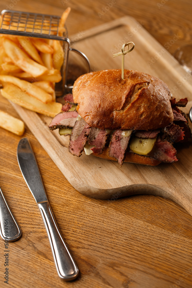 Pastrami sandwich on rye bread served with pickles and mustard sauce. Tasty sandwich with french fries potatoes on dark background.