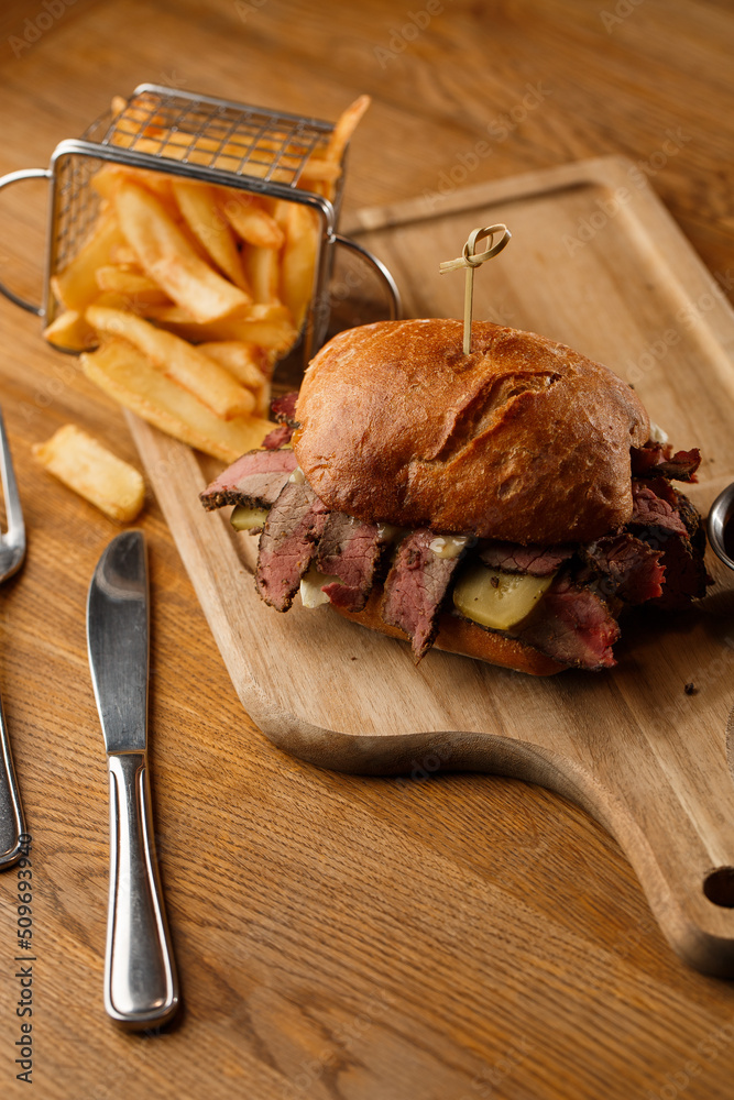 Pastrami sandwich on rye bread served with pickles and mustard sauce. Tasty sandwich with french fries potatoes on dark background.