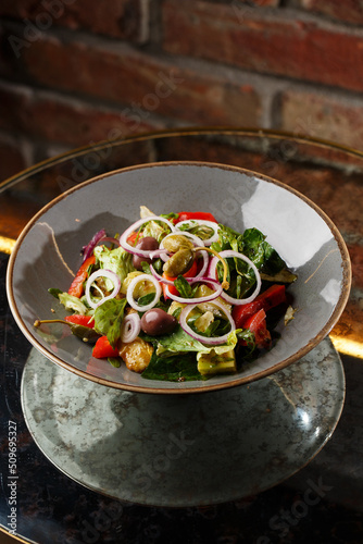 Greek salad with fresh vegetables: tomato, cucumber, red bel pepper, lettuce, onion, olives and cheese. Close-up on a white round plate on a wooden background. Salad menu with ingredients