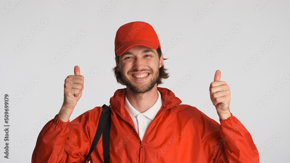 Smiling delivery man dressed in red uniform keeping thumbs up at camera over white background. Postman and express delivery service Smiling delivery man dressed in red uniform keeping thumbs up at camera over white background. Postman and express delivery service