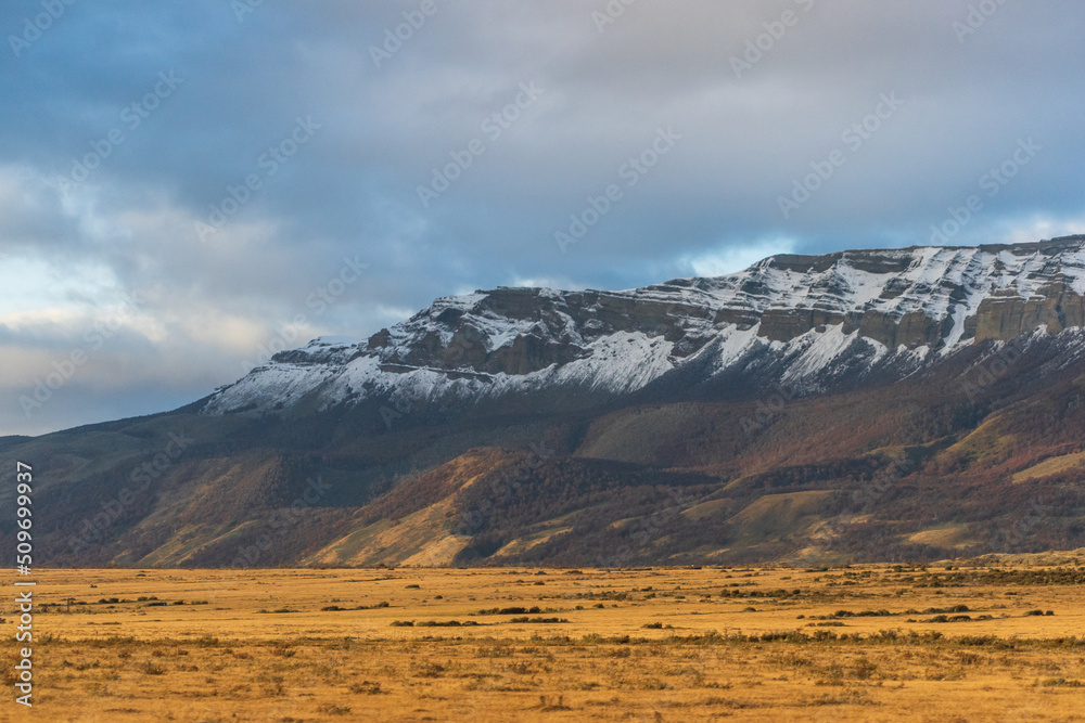 gran cerro nevado con nubes nimbostrato oscuras en atardecer, con el sol de la tarde iluminando campos de colores dorados otoñales  