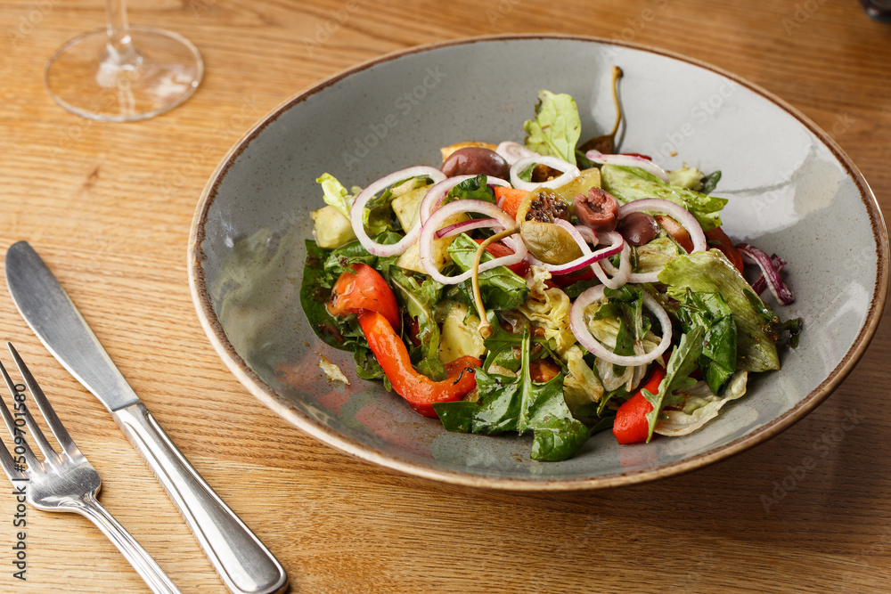 Greek salad with fresh vegetables: tomato, cucumber, red bel pepper, lettuce, onion, olives and cheese. Close-up on a white round plate on a wooden background. Salad menu with ingredients