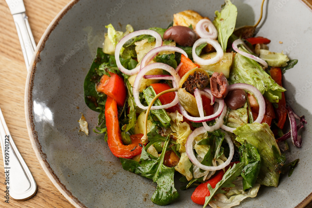 Greek salad with fresh vegetables: tomato, cucumber, red bel pepper, lettuce, onion, olives and cheese. Close-up on a white round plate on a wooden background. Salad menu with ingredients