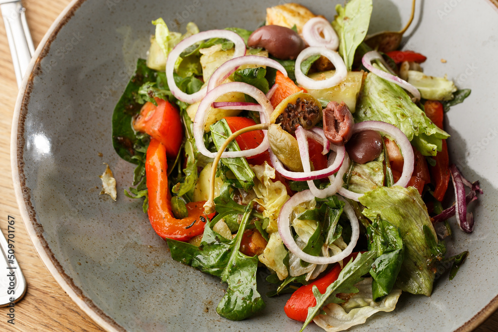 Greek salad with fresh vegetables: tomato, cucumber, red bel pepper, lettuce, onion, olives and cheese. Close-up on a white round plate on a wooden background. Salad menu with ingredients