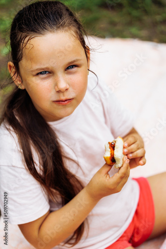 Top view of little hungry girl wth long dark hair wearing pink T-shirt, holding food in hands, biting, sitting in park forest among green trees bushes. Summer, picnic, countryside, nature. Vertical. 