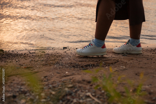 Unrecognizable dark skin woman legs in white boots walking near lake, river or sea side sandy beach. Interracial female on weekends. Water glare and waves. Multicultural female on nature vacation