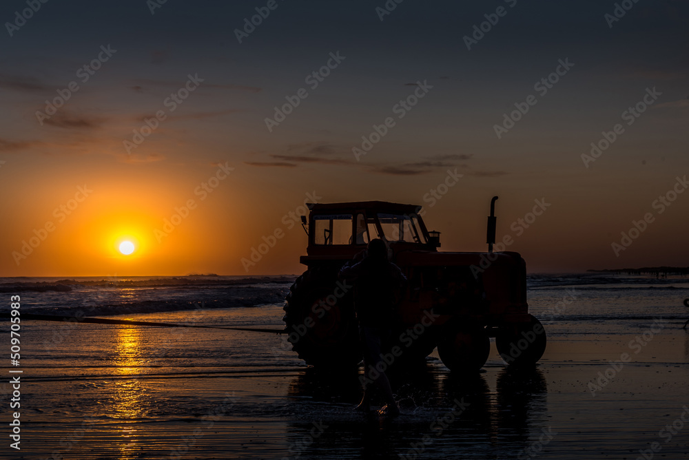 Silhouette of a tractor on the beach at sunset over the sea and reflection on the water