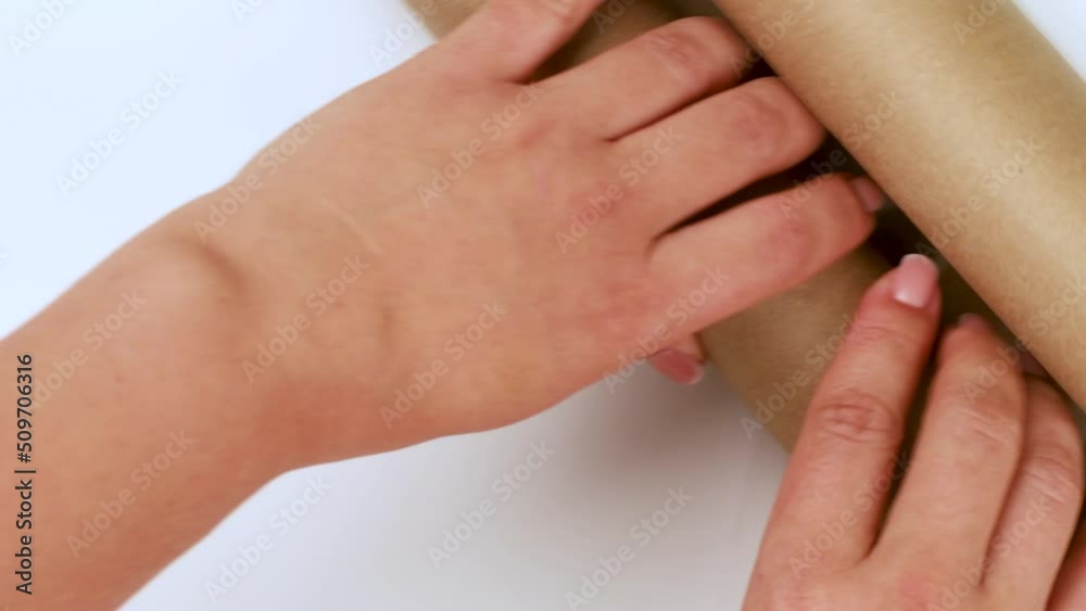 Unrecognizable woman winding up roll of brown kraft paper with manicured hands on white background. Top view. Wrapping presents, handmade, design, art, decorating, eco-friendly products. Close-up. Unrecognizable woman winding up roll of brown kraft paper with manicured hands on white background. Top view. Wrapping presents, handmade, design, art, decorating, eco-friendly products. Close-up.