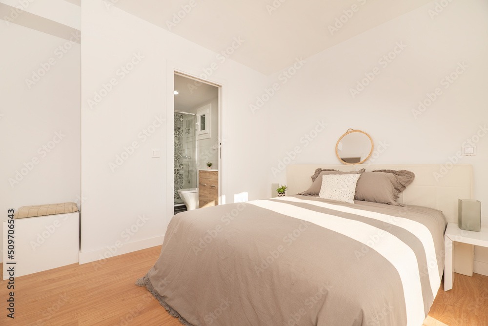 Fototapeta premium Bedroom with double bed with gray bedspread, white furniture and headboard, access to an en-suite toilet and light wooden floor