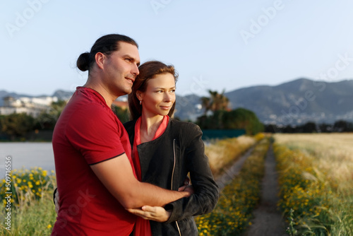 Portrait of lovely middle-aged couple looking aside at copyspace and hugging while standing on grass in summer field.