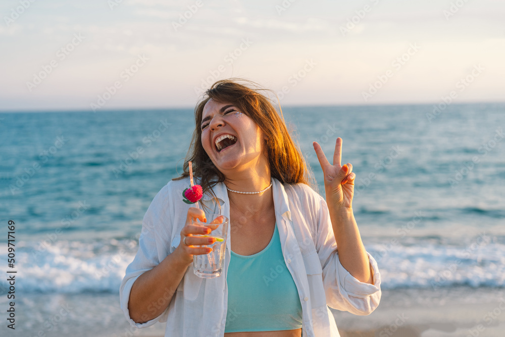 Portrait of a happy young girl with a cocktail in hand on a background of beautiful sea. Girl having fun on summer vacation. The girl shows the peace with two fingers. Freedom and happiness