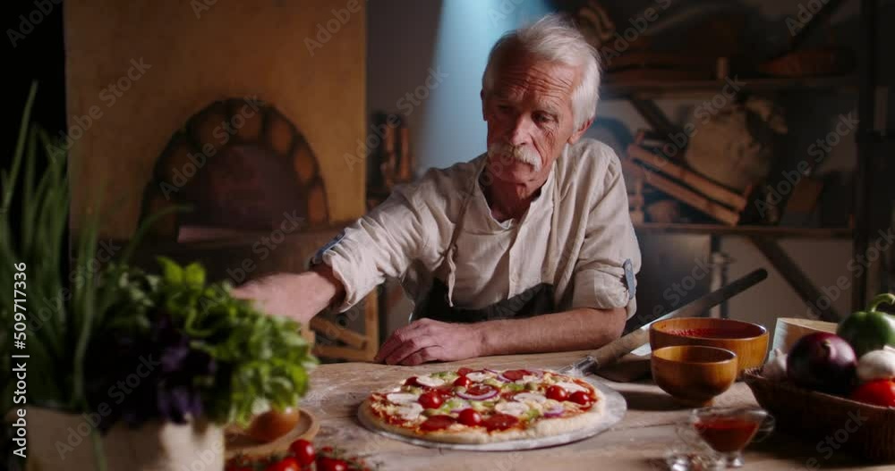 Senior experienced chef is preparing pizza for serving, sprinkling basil leaves. Old cooker using traditional recipe in pizzeria, looking at camera 4k portrait footage Senior experienced chef is preparing pizza for serving, sprinkling basil leaves. Old cooker using traditional recipe in pizzeria, looking at camera 4k portrait footage