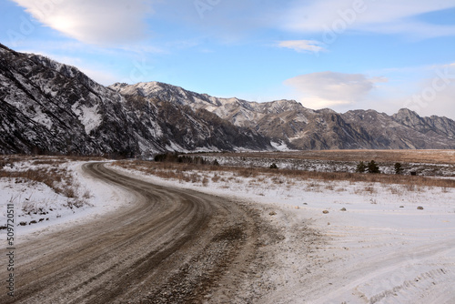 A winding field road running at the foot of snow-capped mountains through a picturesque winter valley.