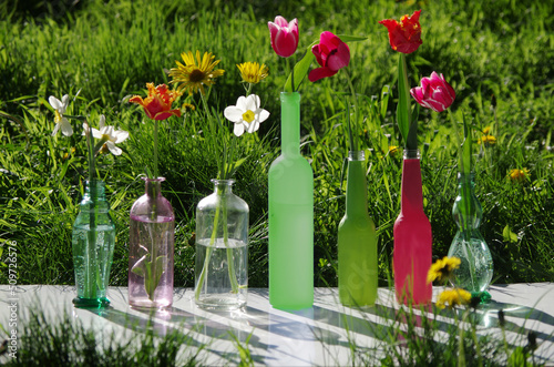Tulips and daffodils in colorful vases and bottles against the background of grass.