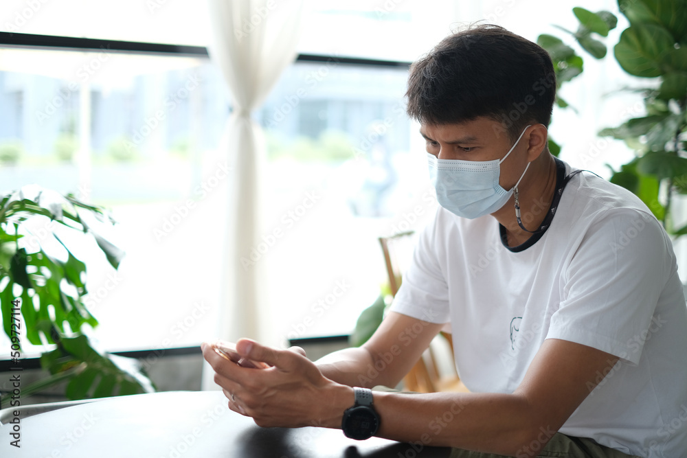 Thai man wearing the medical mask using his mobile phone and reading news about the situation with coronavirus pandemic in the coffee shop. Daily life after covid-19. New normal lifestyle concept.