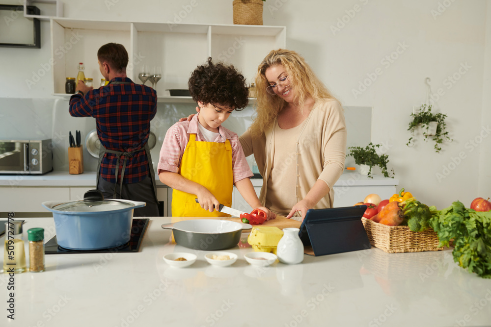 Smiling mother supporting son cutting vegetables for dinner salad