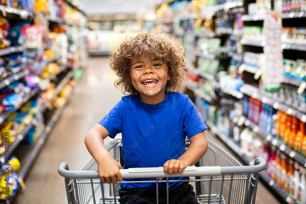 Funny little boy laughing while sitting in a shopping cart during a family trip to the grocery store. He is excited to buy something new	 - obrazy, fototapety, plakaty 
